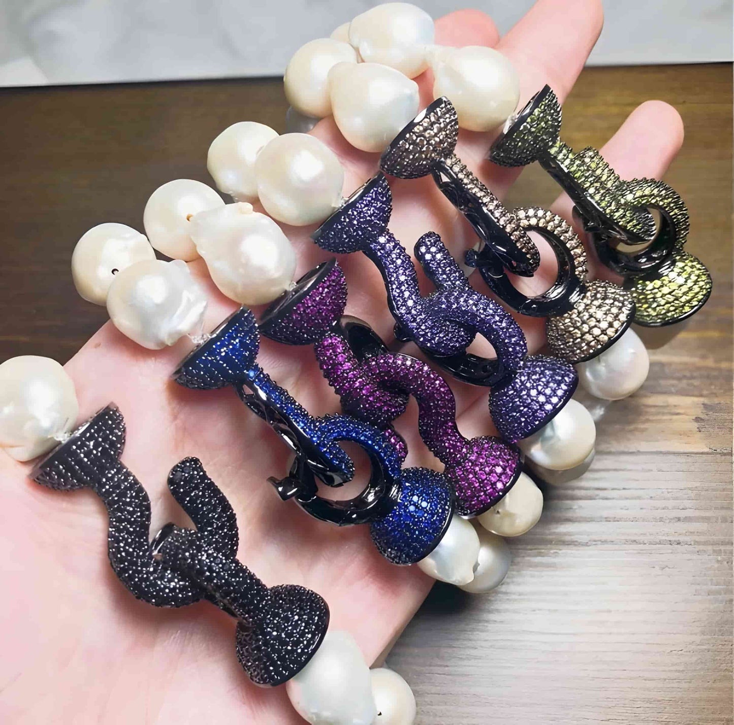 A hand displays five DiamondVeneer Fashion Baroque Fresh Water Pearls w/jeweled Zirconite Bracelets, showcasing large white pearls and jeweled clasps, elegantly arranged on a wooden surface—a must-have for pearl lovers.