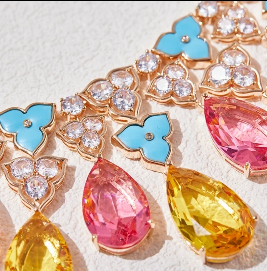 A close-up of the Zirconmania Couture Rain Drop Pear Statement Necklace Earrings Set 840S106, featuring blue clover stones, clear crystals, and large pink and yellow teardrop gems set in gold on a light textured surface.