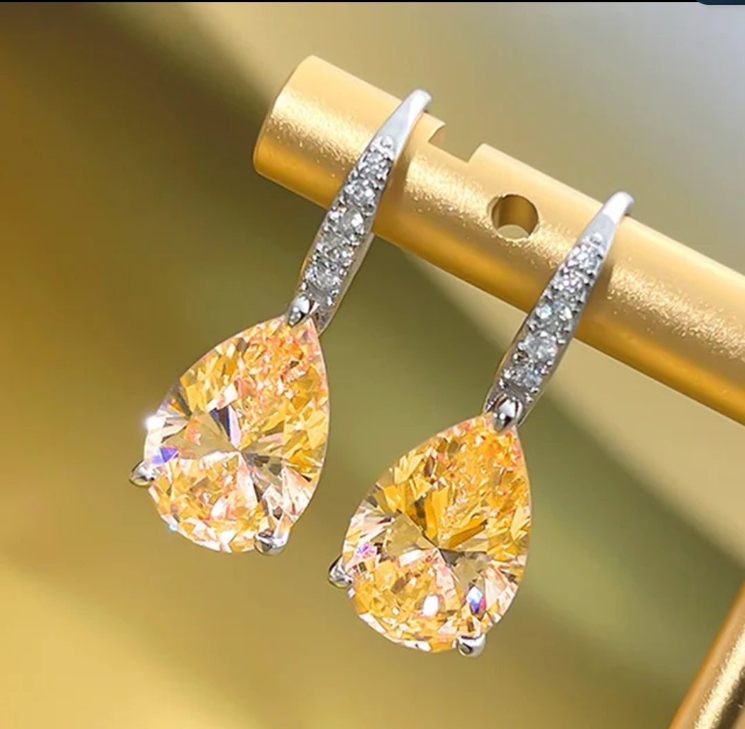 A pair of Cubic Zirconia Pear Earrings Zirconite. 851E2648 by zirconmania fashion jewelry, featuring teardrop yellow stones with silver hooks accented by small white stones, displayed on a gold stand against a blurred background.
