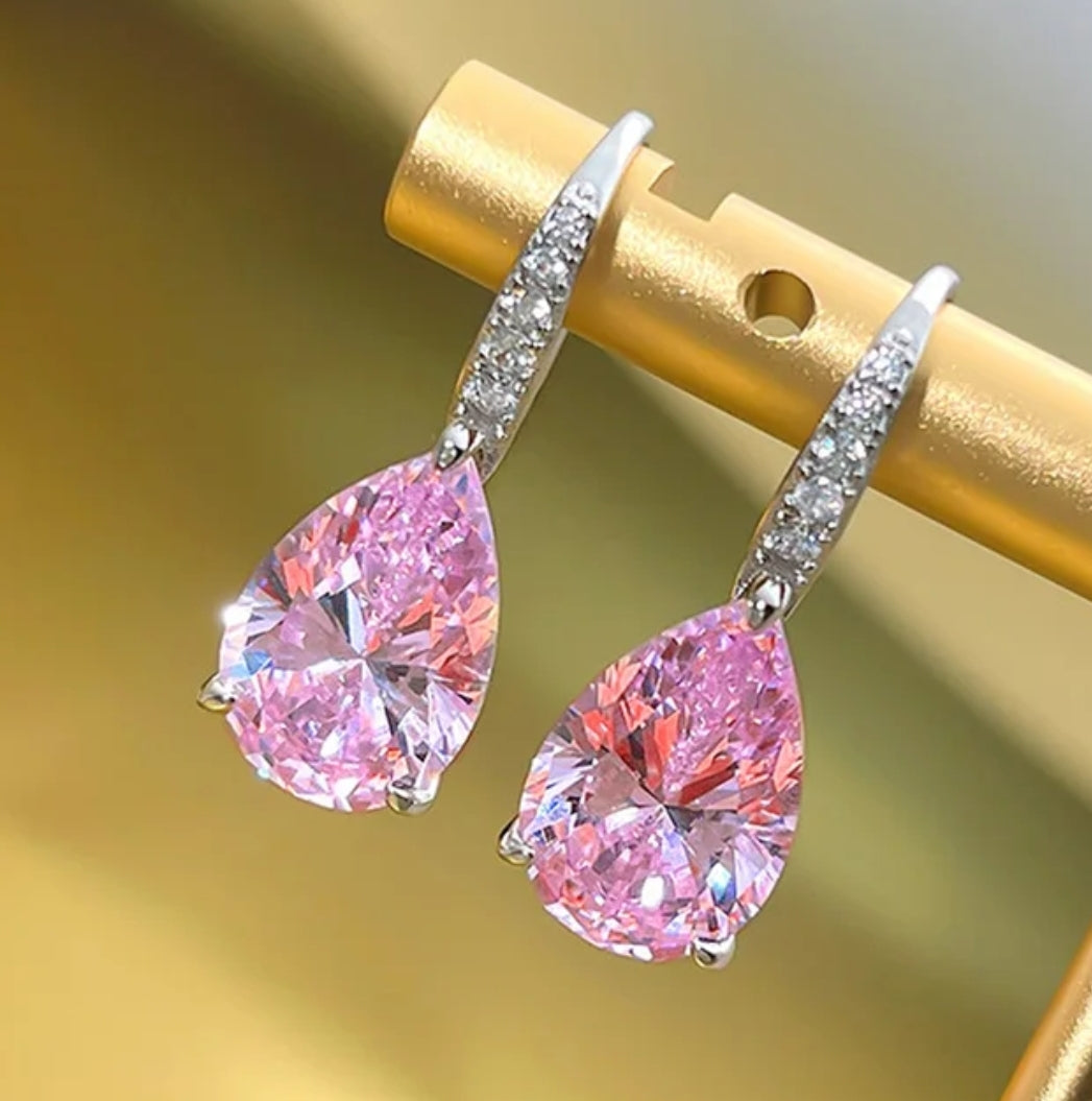 The zirconmania fashion jewelry Cubic Zirconia Pear Earrings (Zirconite 851E2648) feature large pink pear-shaped stones and silver hooks with small Zirconite accents, displayed on a gold rod against a blurred background.