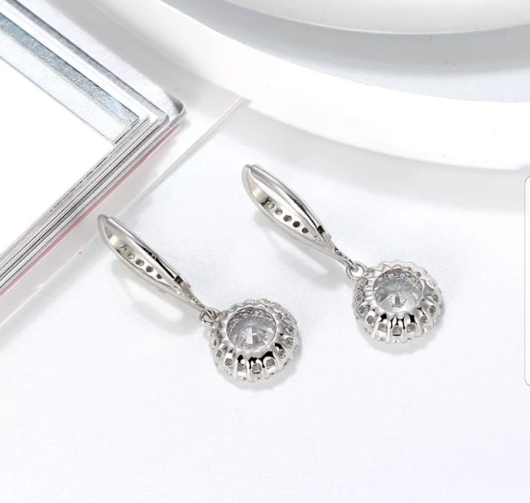 DiamondVeneer Fashions Cubic Zirconia Earrings (635E10687) feature round, clear stones in sterling silver scalloped halo settings, elegantly displayed on a white surface near a book and plate.