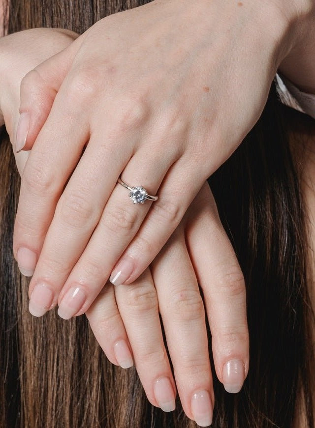 A close-up of two hands with natural nails, one resting on the other. The top hand wears a DiamondVeneer Fashion Round Cubic Zirconia Solitaire Engagement Ring (635R208A). Brown hair appears in the background.
