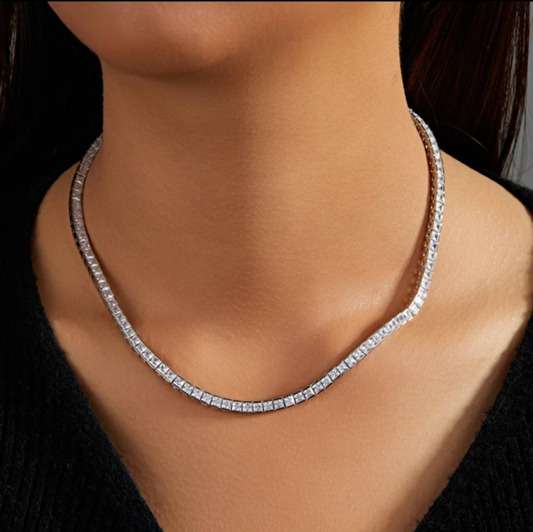 A person wearing the Zirconmania Square Cubic Zirconia CZ Tennis Necklace, style 602N50010 Zirconite, featuring princess cut stones on a thin chain, shown against their bare neck and a black top.