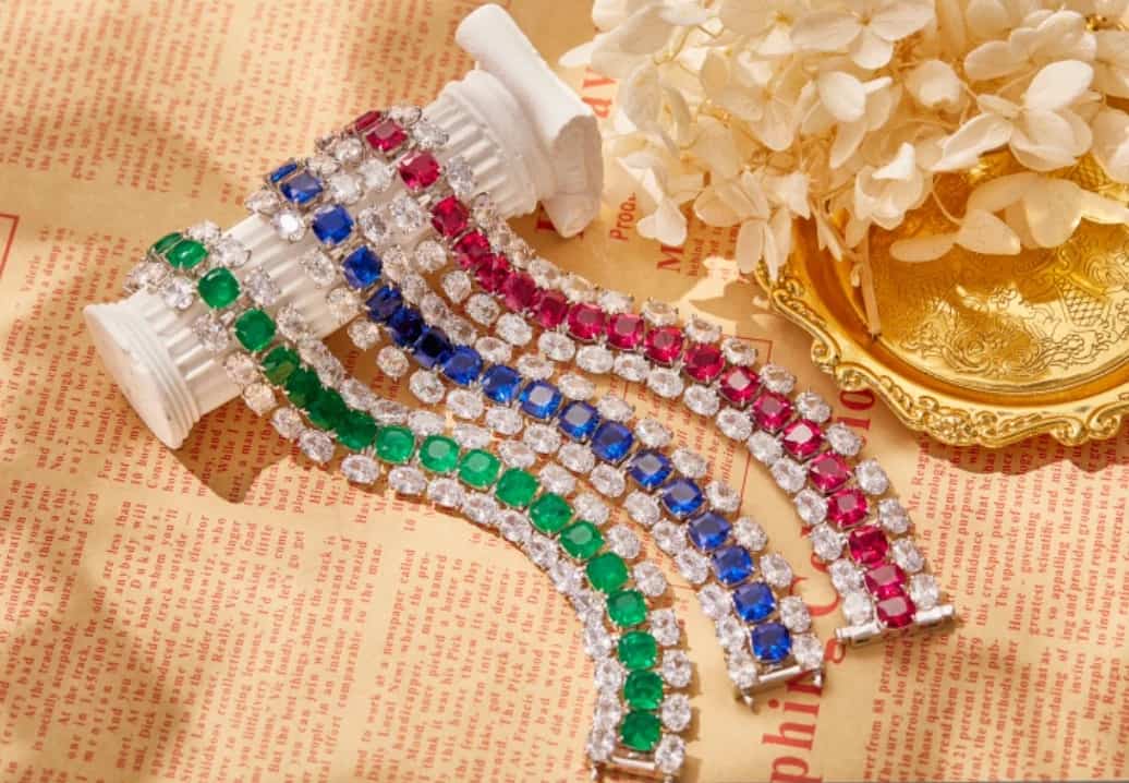 Four Couture Bracelet Zirconite Cubic Zirconia bracelets (840B100) by DiamondVeneer Fashion—green, blue, and pink—are arranged on a white column beside flowers in a golden dish, set against vintage-style paper.