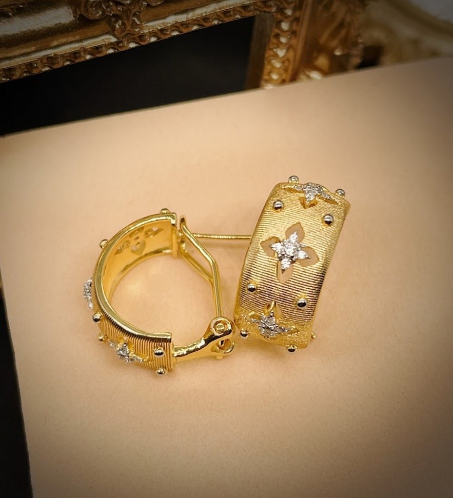 The DiamondVeneer Fashion Italian design satin Hoop Earrings (831E104) feature textured surfaces and clover patterns with diamond-like stones, shown on a beige surface with a gold ornate frame in the background.