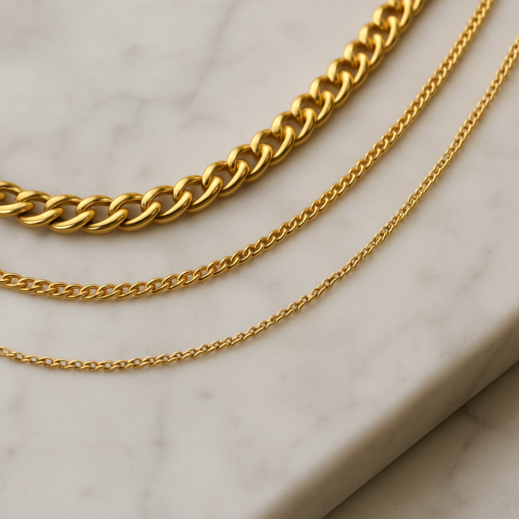 A close‑up of three necklaces of varying thickness neatly arranged on a marble vanity, the thinnest chain closest to the surface and the thickest on top. Alt: arrange necklaces by thickness to prevent tangling