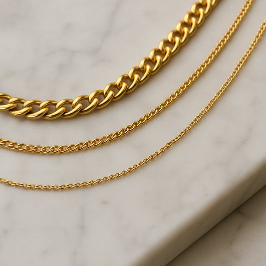 A close‑up of three necklaces of varying thickness neatly arranged on a marble vanity, the thinnest chain closest to the surface and the thickest on top. Alt: arrange necklaces by thickness to prevent tangling