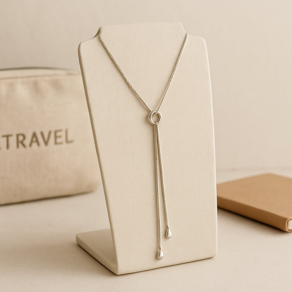 A travel‑ready lariat necklace displayed on a minimalist stand, with a travel pouch and a notebook in the background. Alt: Lariat necklace travel accessory.