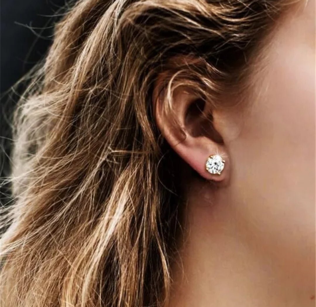 Close-up of a woman's ear adorned with a large, round diamond stud earring, wavy light brown hair framing her ear. Sparkling jewelry detail.