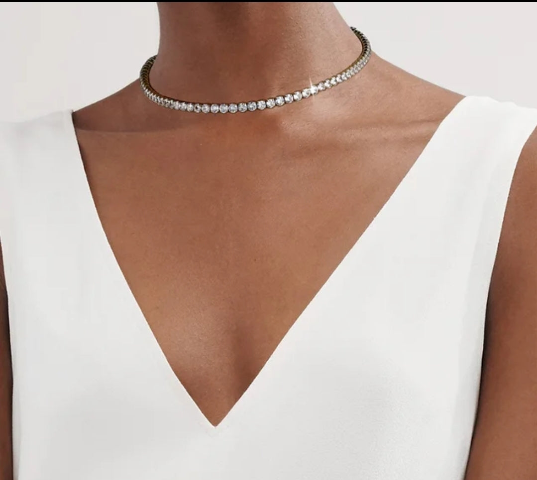 Close-up of a person in a white V-neck top and sparkly gemstone choker necklace, visible from shoulders to chin, on plain background.