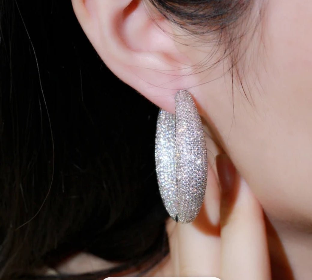 Close-up of a person with large sparkly silver hoop earrings, hand touching ear. Blurred background emphasizes glittery statement earrings.