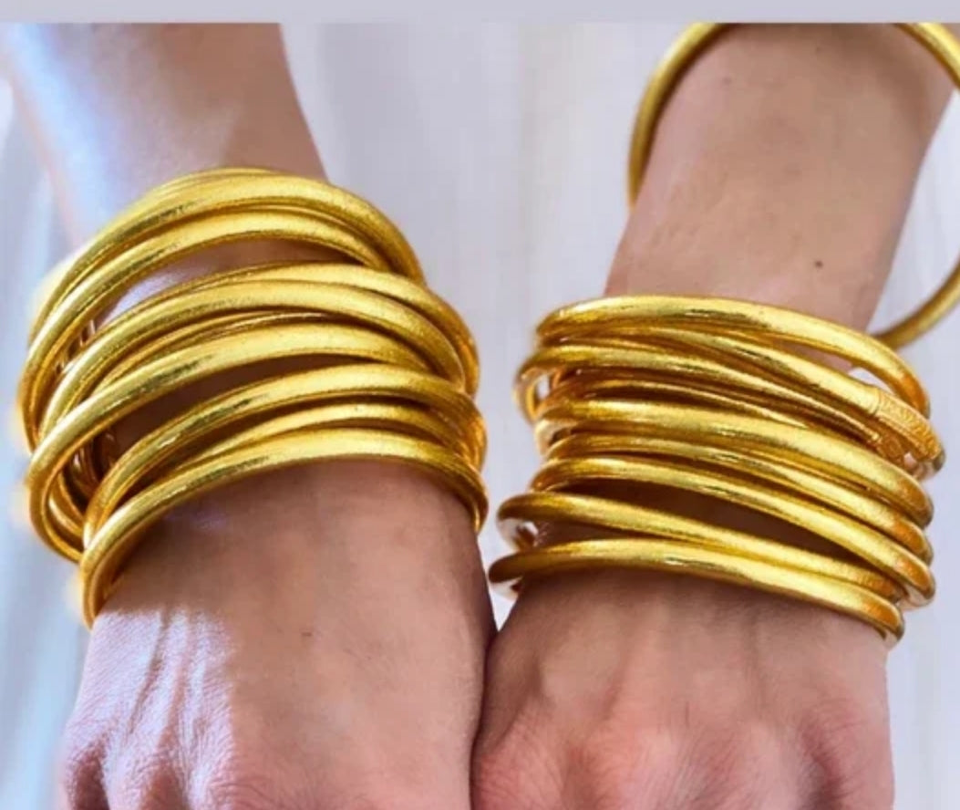 Close-up of two hands wearing multiple Zirconmania Fashion Jewelry Buddhist Gold Silicone Temple Bangle Bracelets (859B100G), showcasing their shiny 18k gold look against a light background.