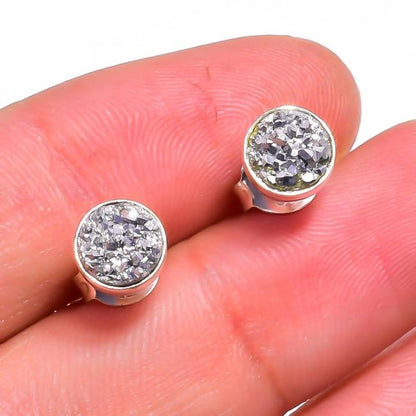 A close-up of two DiamondVeneer Fashion Round Zirconite Cubic Zirconia Sterling Silver Stud Earrings (EBZL) with sparkly, textured surfaces, held between two fingers.