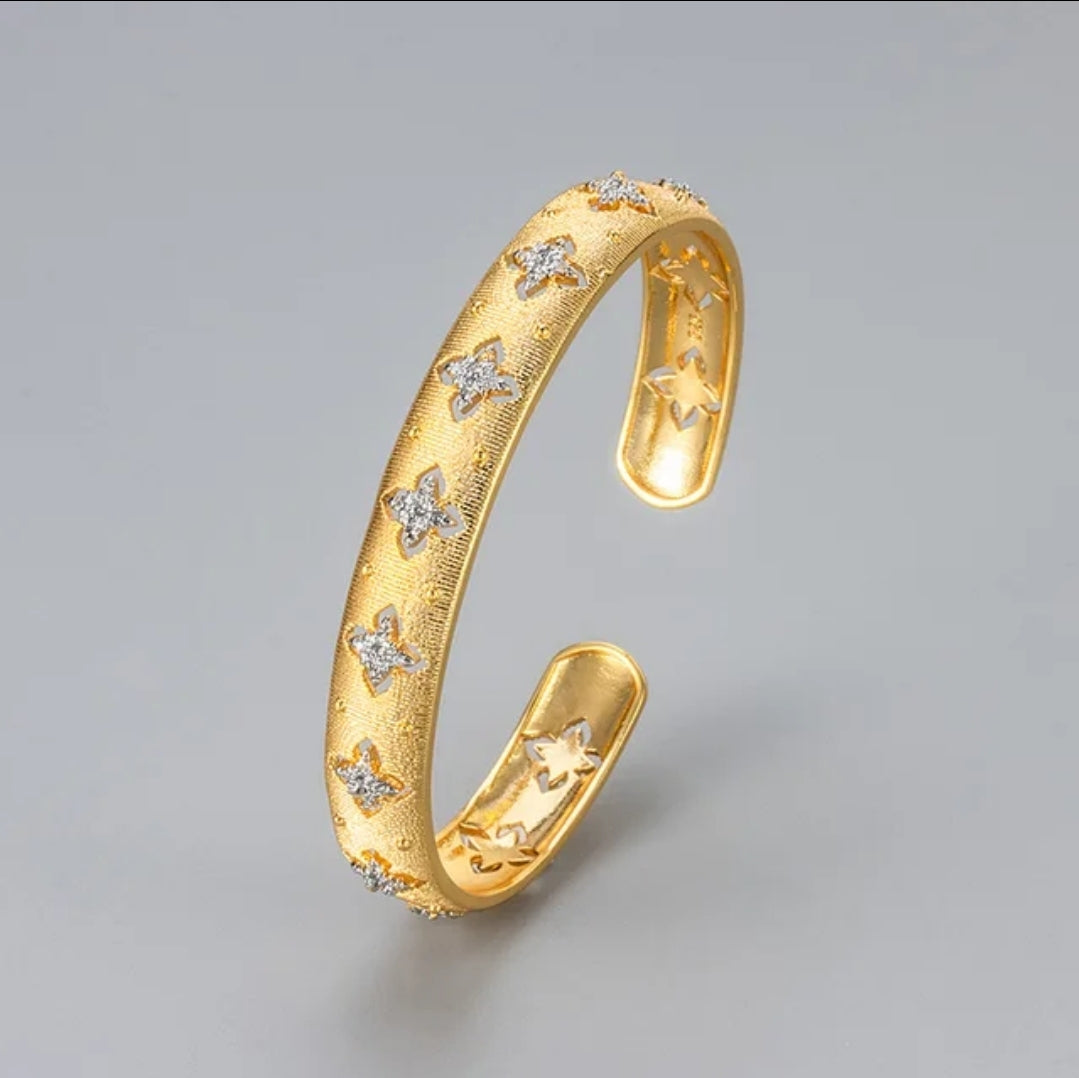 Hand models textured gold open cuff bangle by Zirconmania, featuring a star detail; elegant design on neutral beige background.