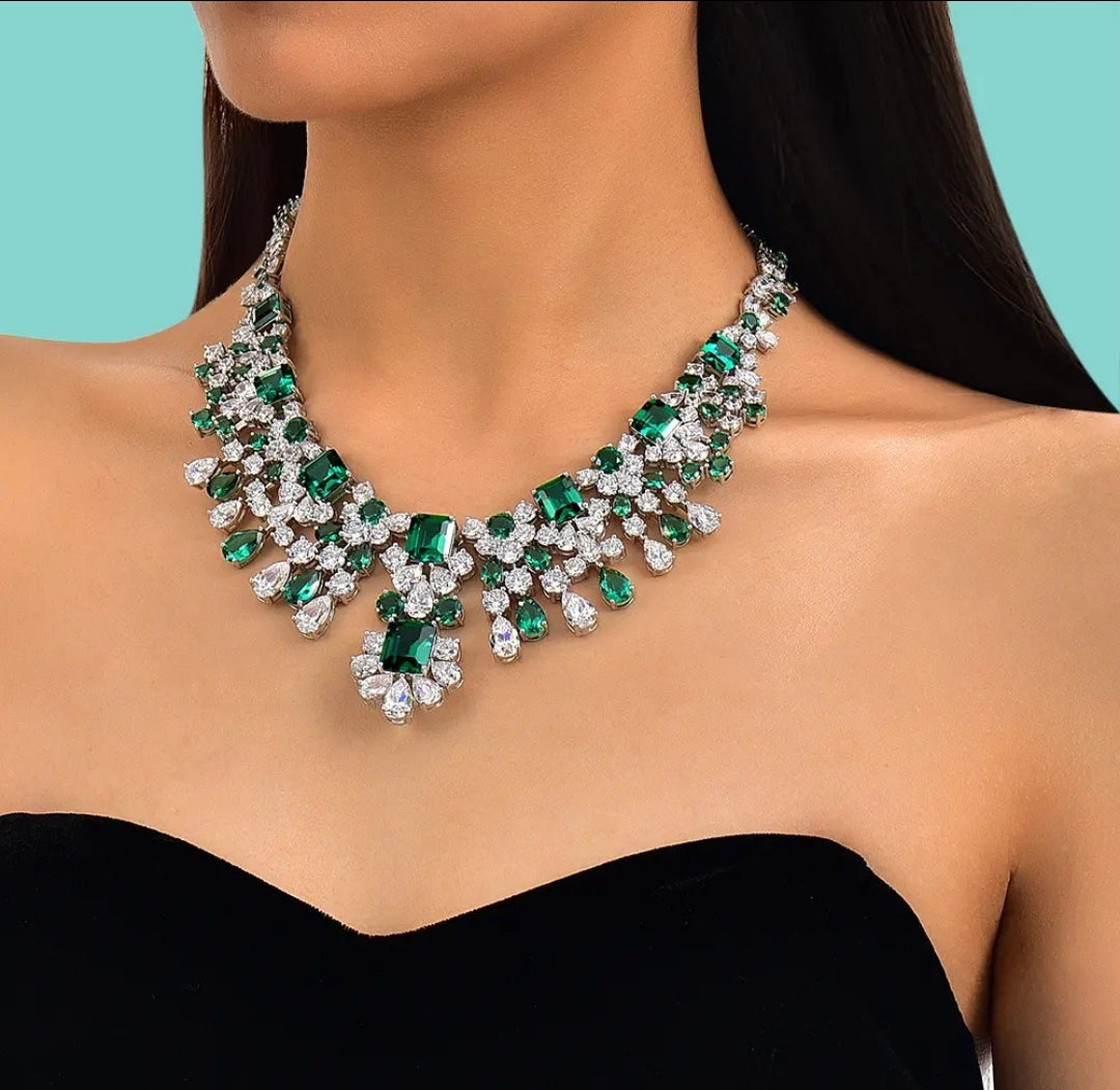 A woman in a strapless black dress models the Zirconmania Couture Statement Luxury Necklace 842N100, featuring large emerald-green and clear gemstones, against a light blue background.