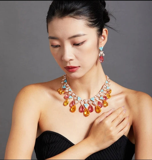 A woman in a strapless black dress gazes down with one hand on her chest, showcasing the DiamondVeneer Fashion Couture Rain Drop Pear Statement Necklace Earrings Set Zirconite 840S106, featuring colorful stones, against a gray background.