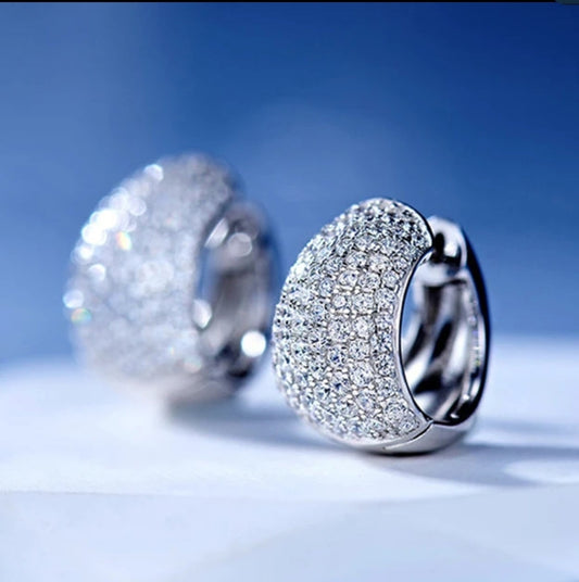 A close-up of DiamondVeneer Fashions Cubic Zirconia tiny Huggies Earrings (Zirconite.851E2445), featuring elegant rhodium plating and pave-set sparkling stones, displayed on a smooth surface with a blue background.