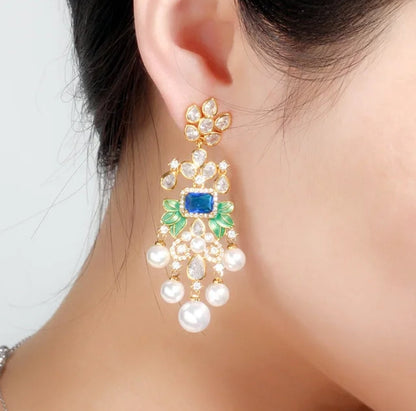 A close-up of a womans ear wearing DiamondVeneer Fashions Whimsical Pearl droplets Earrings Zirconite, featuring blue gemstone, green accents, clear stones, and dangling pearls in a floral chandelier design.