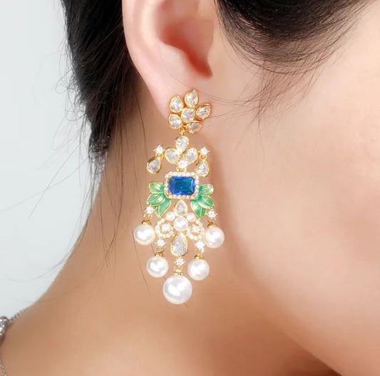 A close-up of a womans ear wearing DiamondVeneer Fashions Whimsical Pearl droplets Earrings Zirconite, featuring blue gemstone, green accents, clear stones, and dangling pearls in a floral chandelier design.