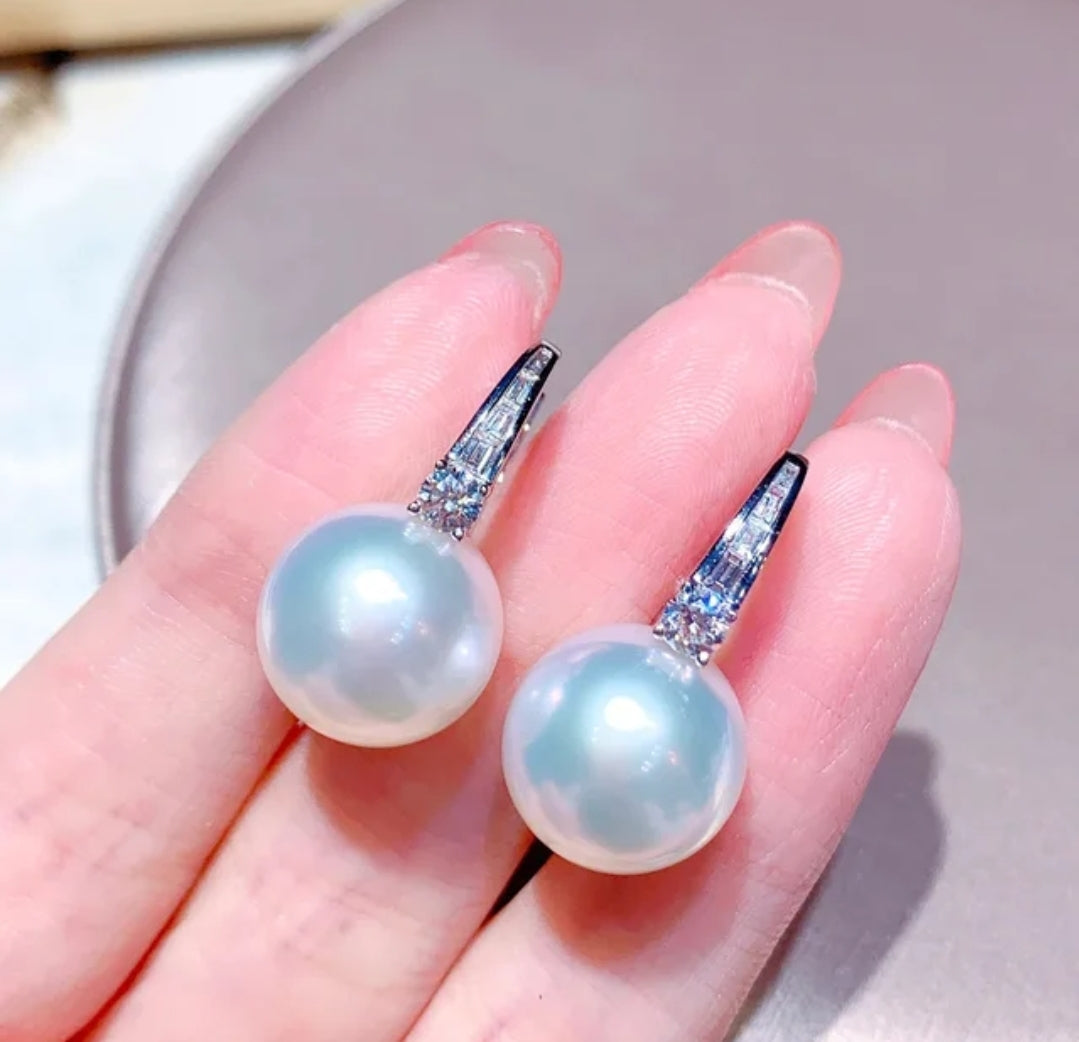 A close-up of a hand holding DiamondVeneer Fashion’s White Pearls Cubic Zirconia Huggies Earrings, featuring large white pearls and sparkling zirconite on silver hooks. 851E2949.
