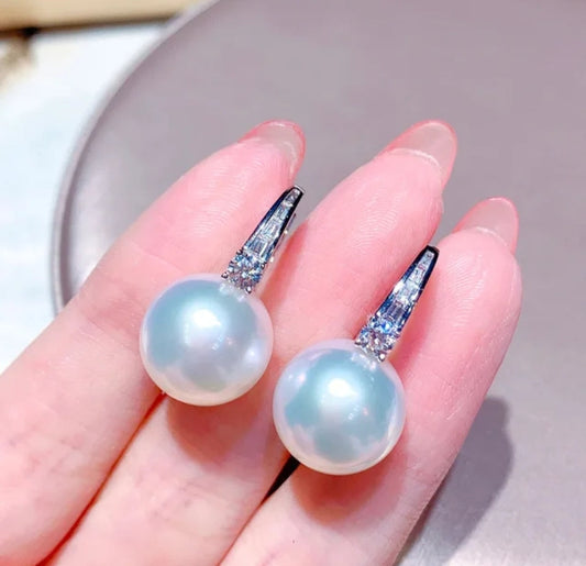 A close-up of a hand holding DiamondVeneer Fashion’s White Pearls Cubic Zirconia Huggies Earrings, featuring large white pearls and sparkling zirconite on silver hooks. 851E2949.