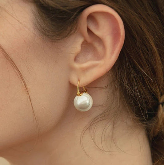 Close-up of a woman wearing DiamondVeneer Fashion’s White Pearls Cubic Zirconia Earrings (Zirconite 826E3092): gold hoops with large round white pearl charms. Her brown hair is tied back against a softly blurred background.