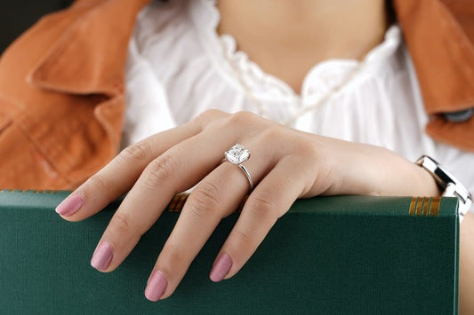 A hand with light pink nail polish wears the DiamondVeneer Fashion Cushion Diamond Veneer Cubic Zirconia 14K Gold Solitaire Ring (635R208K), resting on a green book. The person is dressed in a white blouse and brown jacket.