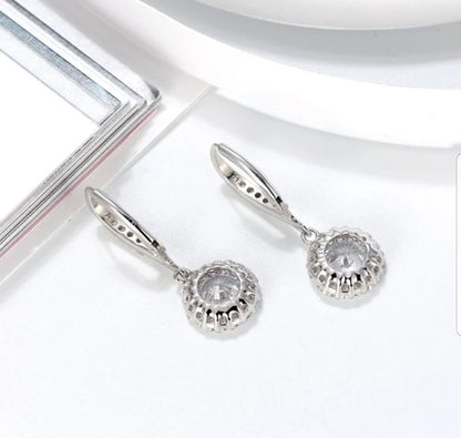 DiamondVeneer Fashions Cubic Zirconia Earrings (635E10687) feature round, clear stones in sterling silver scalloped halo settings, elegantly displayed on a white surface near a book and plate.