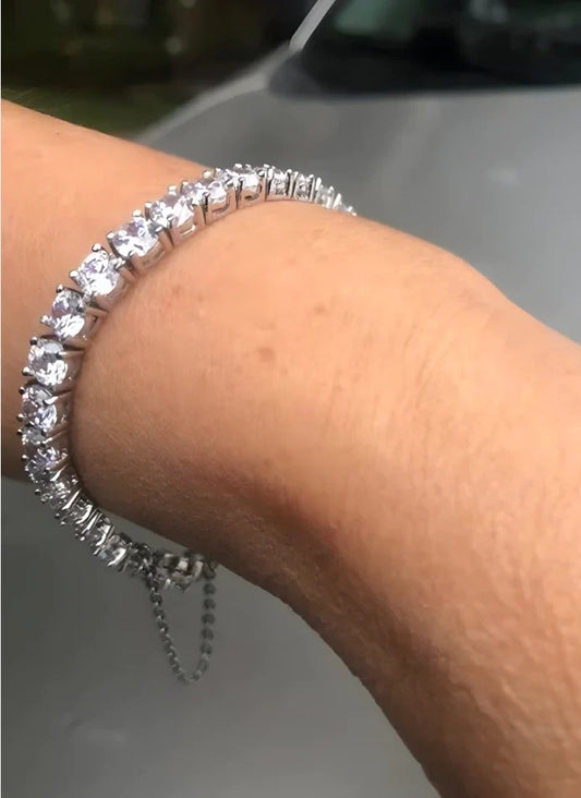 A close-up shows a persons wrist adorned with the DiamondVeneer Fashion Cubic Zirconia Tennis Bracelet with Safety Chain 698Bcast-Zirconite, featuring round, clear stones in a row against a blurred gray background.