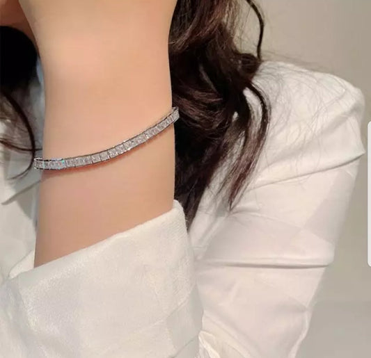 A person in a white long-sleeve shirt wears the DiamondVeneer Fashion Princess Cut Square Zirconite CZ Tennis Bracelet (B602B30017) on their wrist, with their long dark hair partially visible.