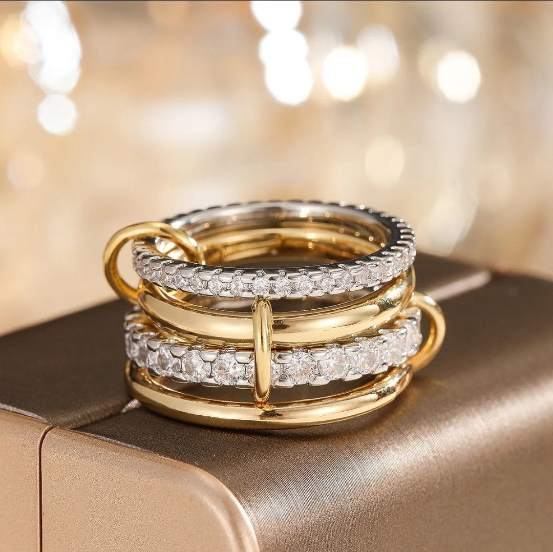 The Zirconmania Fashion Jewelry Ring Interlinked Stacked Zirconite Rings Set (858R100) features gold and silver bands with sparkling pavé, shown atop a metallic gold surface—an elegant choice for luxury travel wear.