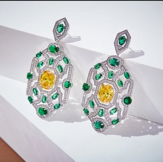 DiamondVeneer Fashion’s Italian inspired Earrings Design Zirconite 840E107 feature large yellow center stones, surrounded by green gemstones and silver accents, displayed on a white geometric surface.
