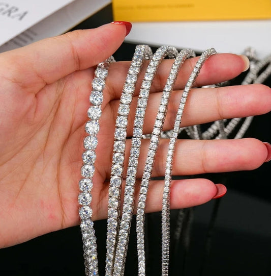 A hand with red-painted nails holds several sparkling silver tennis necklaces, including a DiamondVeneer Fashion Moissanite Lab Created Diamond Tennis Bracelet (850B), all encrusted with round and square-cut clear gemstones against a dark background.