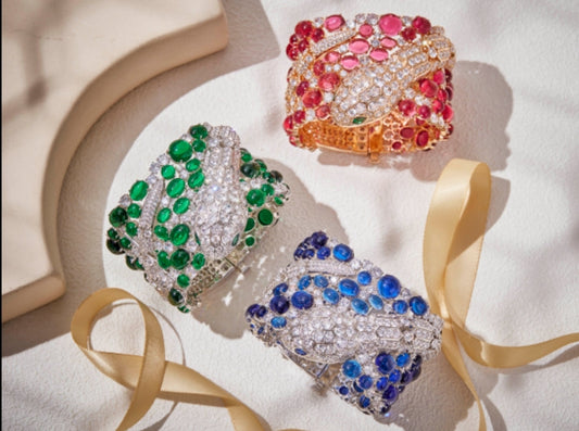 Three jeweled bracelets, including the DiamondVeneer Fashion Luxurious Cabochon Bangle Zirconite 840B300 shaped like coiled snakes and adorned with green, blue, and pink stones, are arranged on a light surface with a gold ribbon nearby.