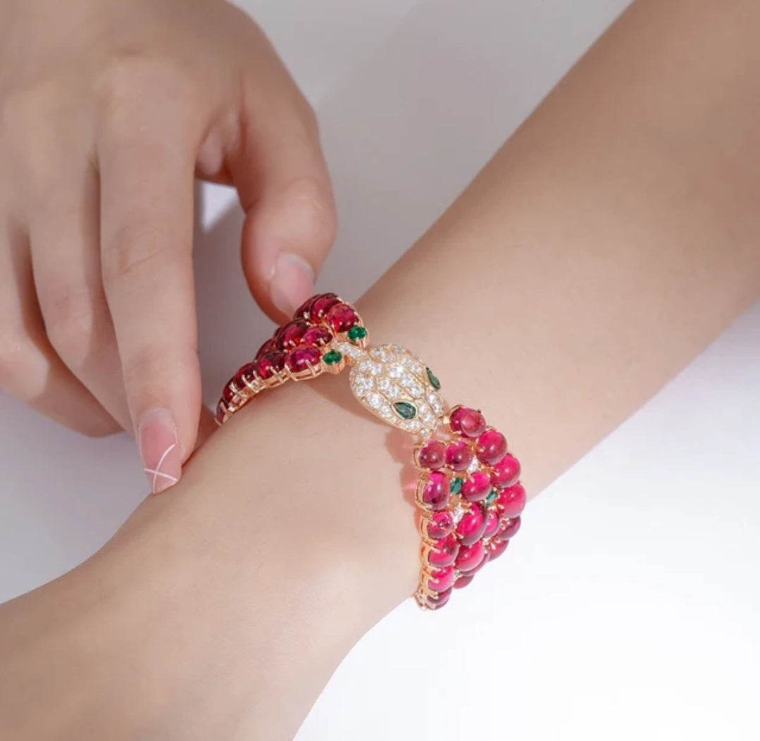 A hand adjusts the DiamondVeneer Fashion Luxury Ruby Cabochon Snake Bracelet Bangle zirconite 840B—a gold snake bangle with red cabochon gemstones, white stones, and green eyes—showcasing exquisite fashion jewelry.