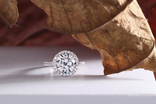 A DiamondVeneer Fashion Round Cubic Zirconia Engagement Halo Ring (635R2054) sits on a white surface, partially covered by a large, dried brown leaf.