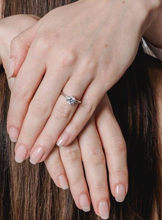 A close-up of two hands with natural nails, one resting on the other. The top hand wears a DiamondVeneer Fashion Round Cubic Zirconia Solitaire Engagement Ring (635R208A). Brown hair appears in the background.