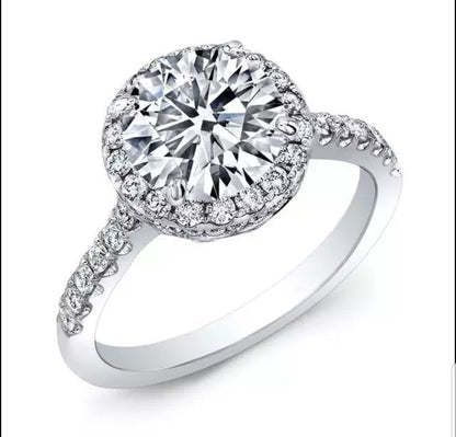 The DiamondVeneer Fashion Round Cubic Zirconia Halo Engagement Ring (800R70/2) features a striking center stone, haloed by smaller gems and accented with extra stones along the white gold band for added brilliance.