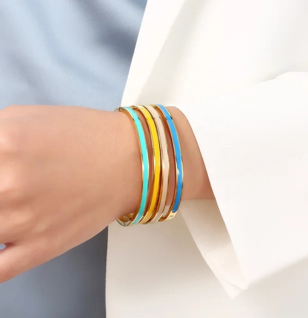 A person in a white blazer and blue top wears four stackable bangles, including the DiamondVeneer Fashion Stackable Enamel Bracelet Bangle Zirconite. 852B100, in gold, blue, and turquoise enamel finishes on their wrist.