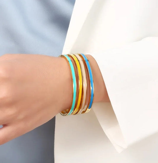A person in a white blazer and blue top wears four stackable bangles, including the DiamondVeneer Fashion Stackable Enamel Bracelet Bangle Zirconite. 852B100, in gold, blue, and turquoise enamel finishes on their wrist.