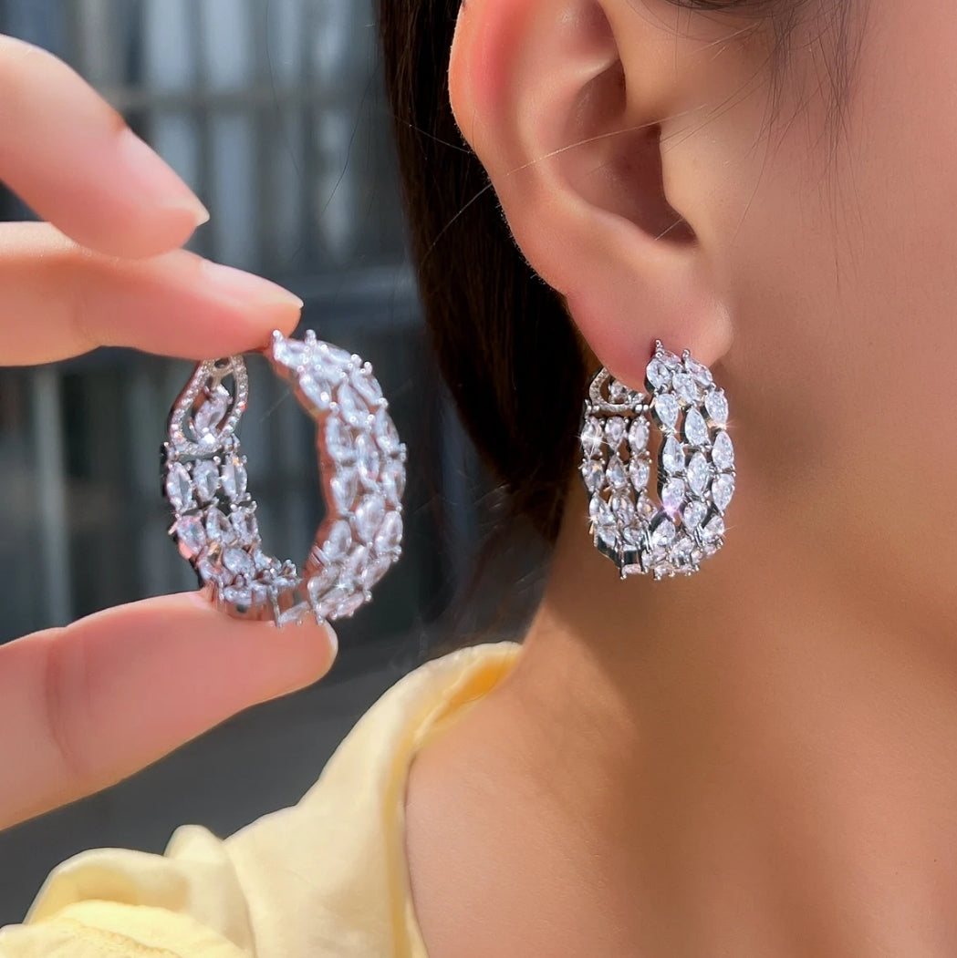 A person showcases Zirconmania Fashion Jewelry’s Hoop Earrings – Cubic Zirconia Classic Cluster Earrings (826E07), holding one near their ear as its sparkling marquise and round stones draw focus against a blurred background.