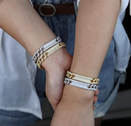 Two arms adorned with chunky chain bracelets, including the DiamondVeneer Fashion Fully Pave set Zirconite Cubic Zirconia ID Curved Link Bracelet.806B100, in gold and silver. The standout pieces complement denim shorts, a white shirt, and a brown belt.