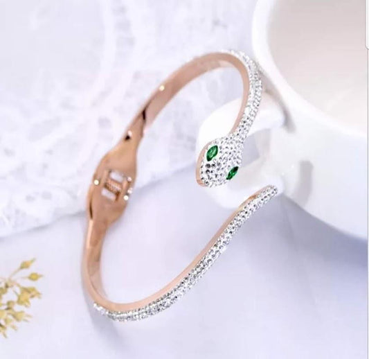 The DiamondVeneer Fashion Face-off Snake Crystals Stainless Steel hinged Bangle Bracelet (655B4), featuring white stones and green gemstone eyes, is elegantly wrapped around a white teacup handle on a pristine surface.