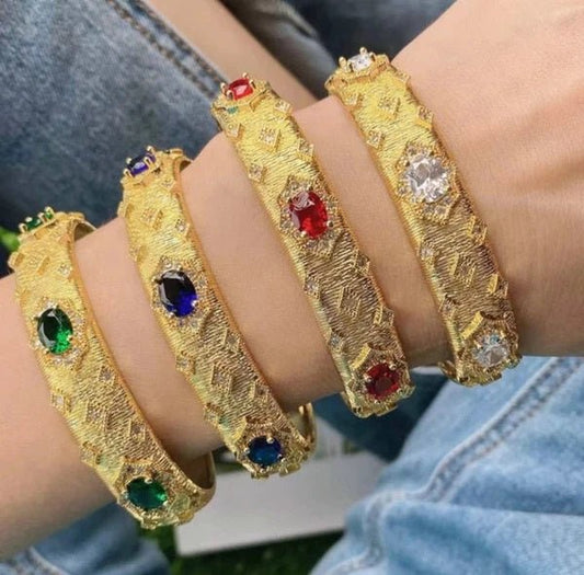 Four DiamondVeneer Fashion Zirconite satin gold open Jeweled Bangle Bracelets (835B100), each featuring a unique gemstone—green, blue, red, and clear cubic zirconia—worn together with denim jeans visible in the background.