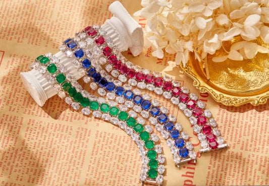 Four Couture Bracelet Zirconite Cubic Zirconia bracelets (840B100) by DiamondVeneer Fashion—green, blue, and pink—are arranged on a white column beside flowers in a golden dish, set against vintage-style paper.