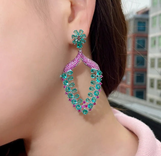 A close-up of a person wearing DiamondVeneer Fashions Zirconite Cubic Zirconia long color Earrings (826E08) with green and pink gemstones in a floral, cascading design, set against a blurred cityscape.
