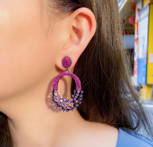 A close-up of a womans ear wearing DiamondVeneer Fashion Chandeliers Earrings Zirconite Cubic Zirconia 826E20, featuring large purple chandelier design with small purple and pink stones and a black rhodium electroplate finish.