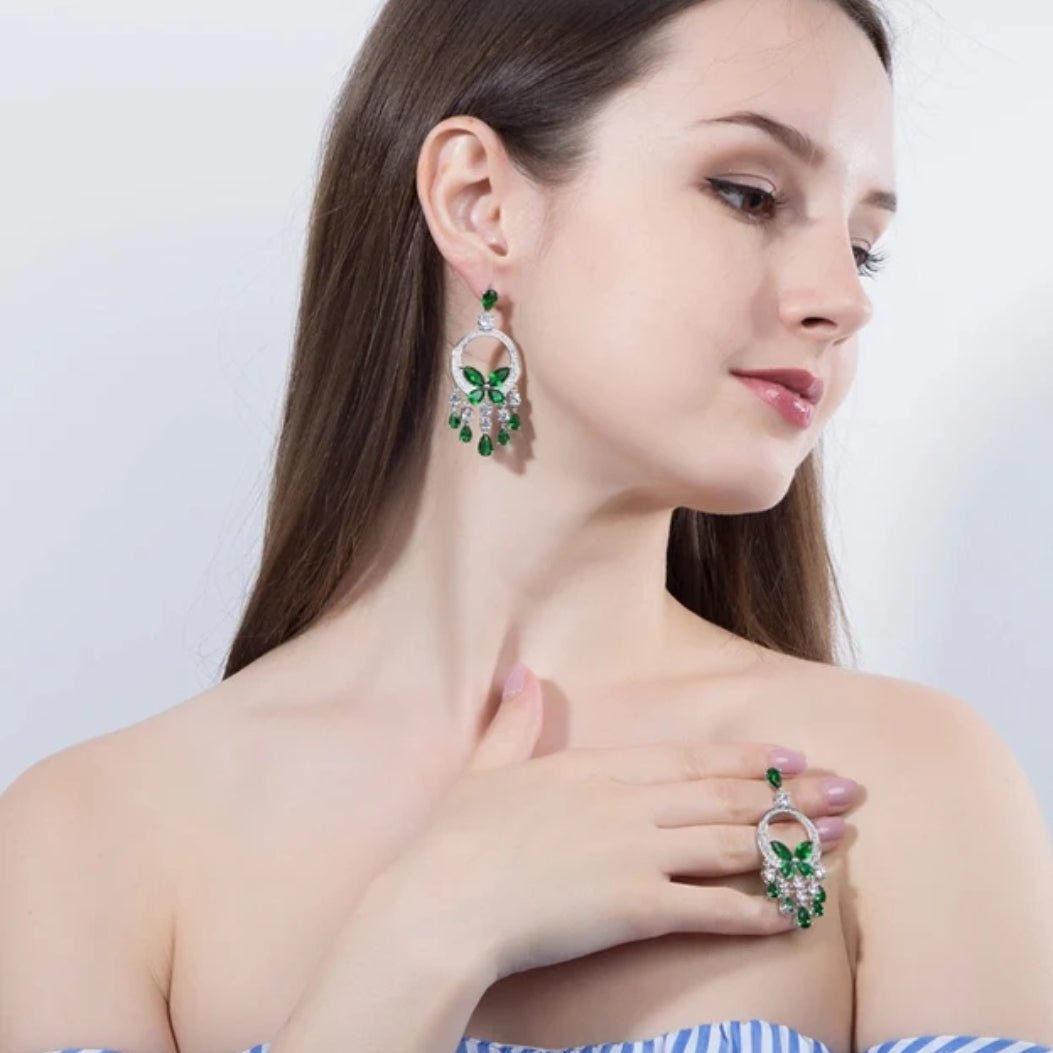 A woman with long brown hair in a white off-shoulder top showcases DiamondVeneer Fashion’s Butterfly Chandeliers Earrings Zirconite Cubic Zirconia 826E01 and a silver-green statement ring, both matching as she touches her collarbone.