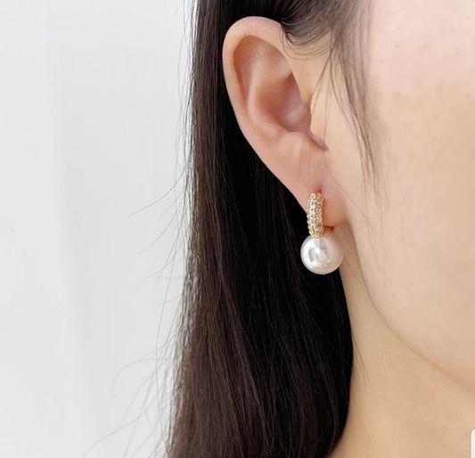 Close-up of a person’s ear wearing DiamondVeneer Fashion Pearl Huggies Earrings Zirconite Cubic Zirconia (836E100), featuring gold texture and large white drop, with straight dark hair visible against a plain white background.