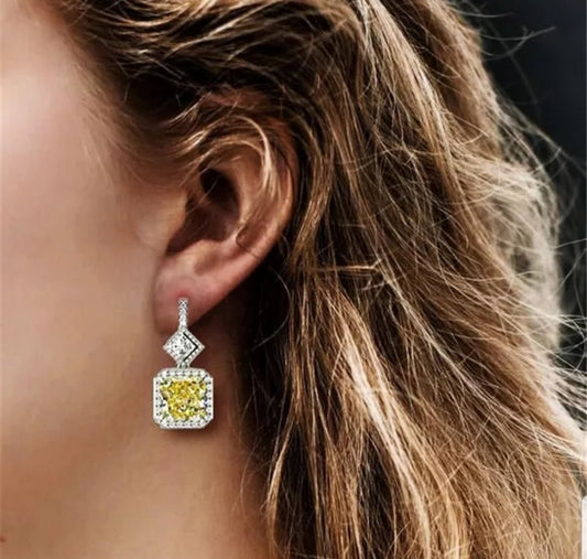 Close-up of a womans ear with wavy light brown hair, wearing DiamondVeneer Fashion Earrings Geometric Diamond Veneer Cubic Zirconia 830E1029, featuring a bright yellow gemstone and smaller white stones in a square setting.
